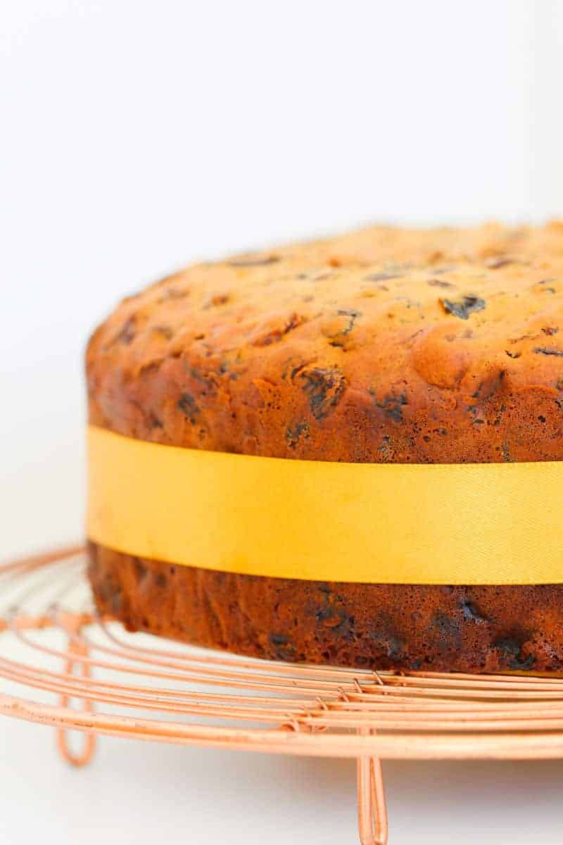 A fruit cake with a gold ribbon around it on a copper wire tray.