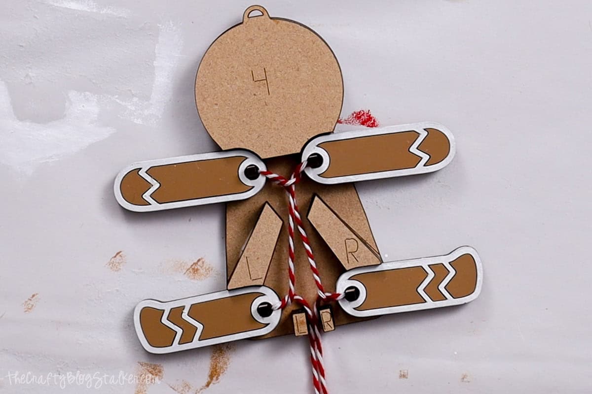 Completed pull-string mechanism on the back of a gingerbread man ornament, with arms and legs securely tied using red and white twine for interactive movement.