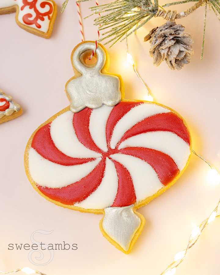 A cookie decorated to look like an ornament with a red and white swirl icing pattern. The cookie has a string through a hole so that it can be hung on a tree. The cookie is surrounded by glowing fairy lights and evergreens.