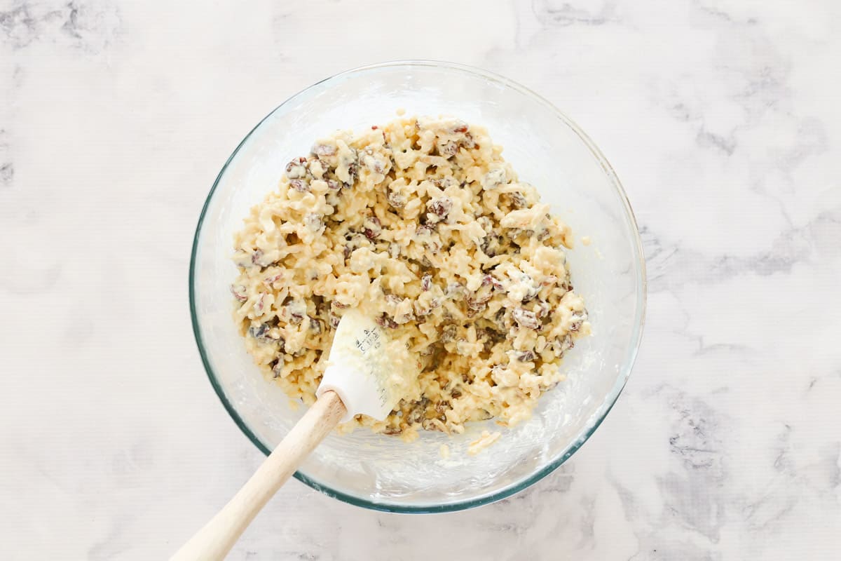 Rice bubbles, dried cranberries, sultanas, almonds, coconut and melted white chocolate mixed with a spatula in a bowl.
