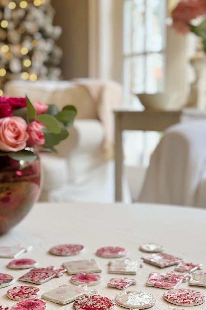 A cozy living room with a Christmas tree in the background. The foreground showcases a table adorned with decorative red and white ornaments, snowglobe cookies, and a vase of pink roses, creating a festive holiday atmosphere.