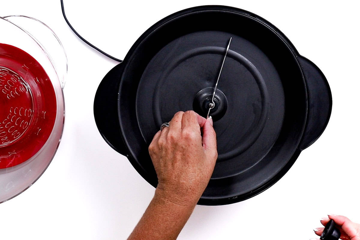 A hand removing the stir rod from the black base of a WestBend Stir Crazy popcorn machine, with the red lid and clear bowl placed to the side.