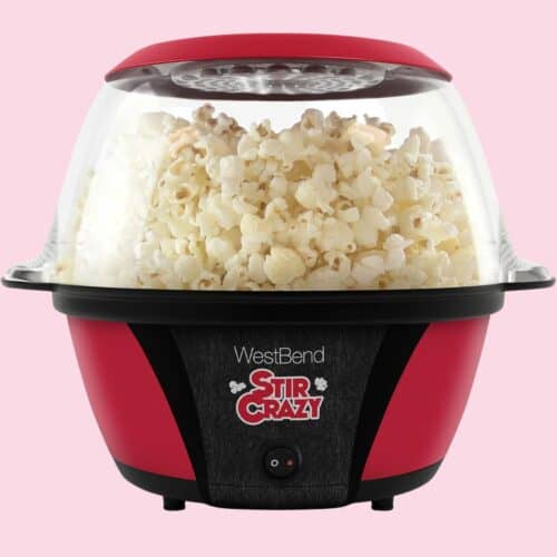 A red and black WestBend Stir Crazy popcorn machine filled with freshly popped popcorn, featuring a clear bowl lid and pink background.