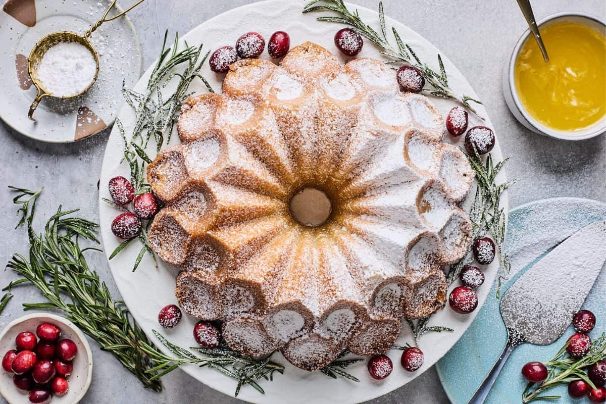 Buttered rum eggnog pound cake on a white plate on the table with cranberries, rosemary, powdered sugar, and a small bowl of sauce.