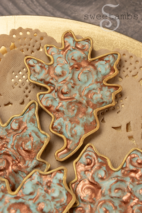 An oak leaf shaped cookie decorated with an embossed filigree design and a faux oxidized copper finish. The cookies are on a kraft paper doily on a gold platter on a wooden table.