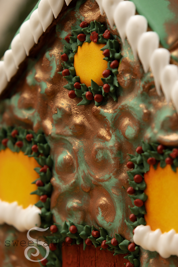A close up of the gingerbread house decorated to look like oxidized copper with an embossed filigree design. The glowing windows are surrounded by royal icing evergreens with berries and there is royal icing snow and icicles on the window sills and along the roof.