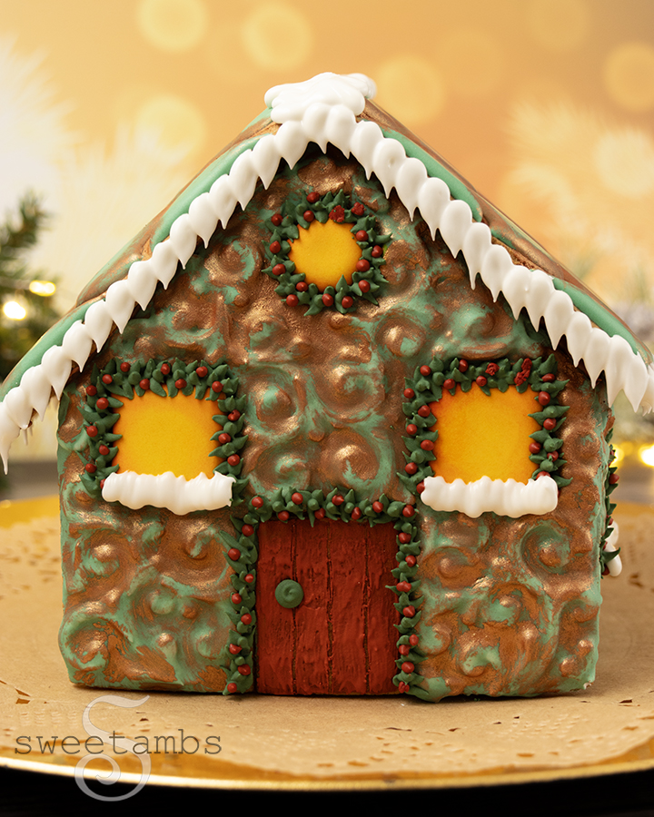 A front view of the gingerbread house decorated to look like oxidized copper and an embossed filigree design. The windows are painted with yellow and orange to look like they're glowing. The gingerbread house is on a kraft paper doily on a gold plate with bokeh lights, fairy lights, and evergreen boughs in the background.