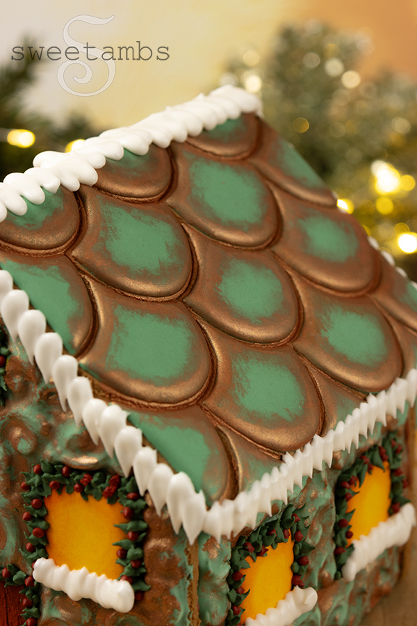 A view of the roof of the gingerbread house. The roof has a scalloped design that is decorated to look like oxidized copper. It is lined with royal icing snow and icicles.