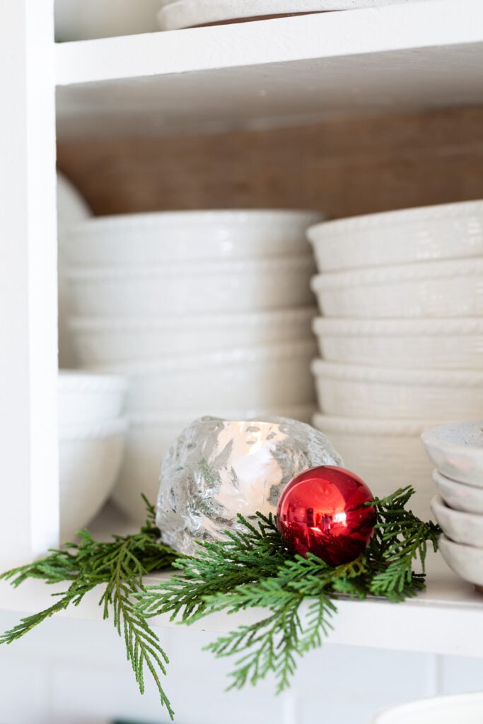 Kosta Boda snowball votive holder on an open kitchen shelf with greenery around it and a red ball ornament.