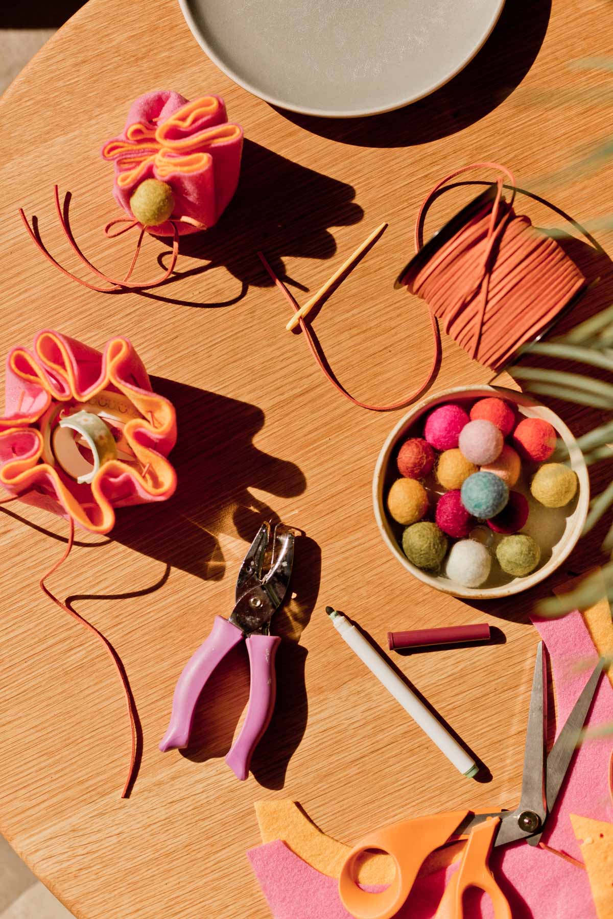 craft supplies styled on wood table, with felt balls, scissors, cording, and more 