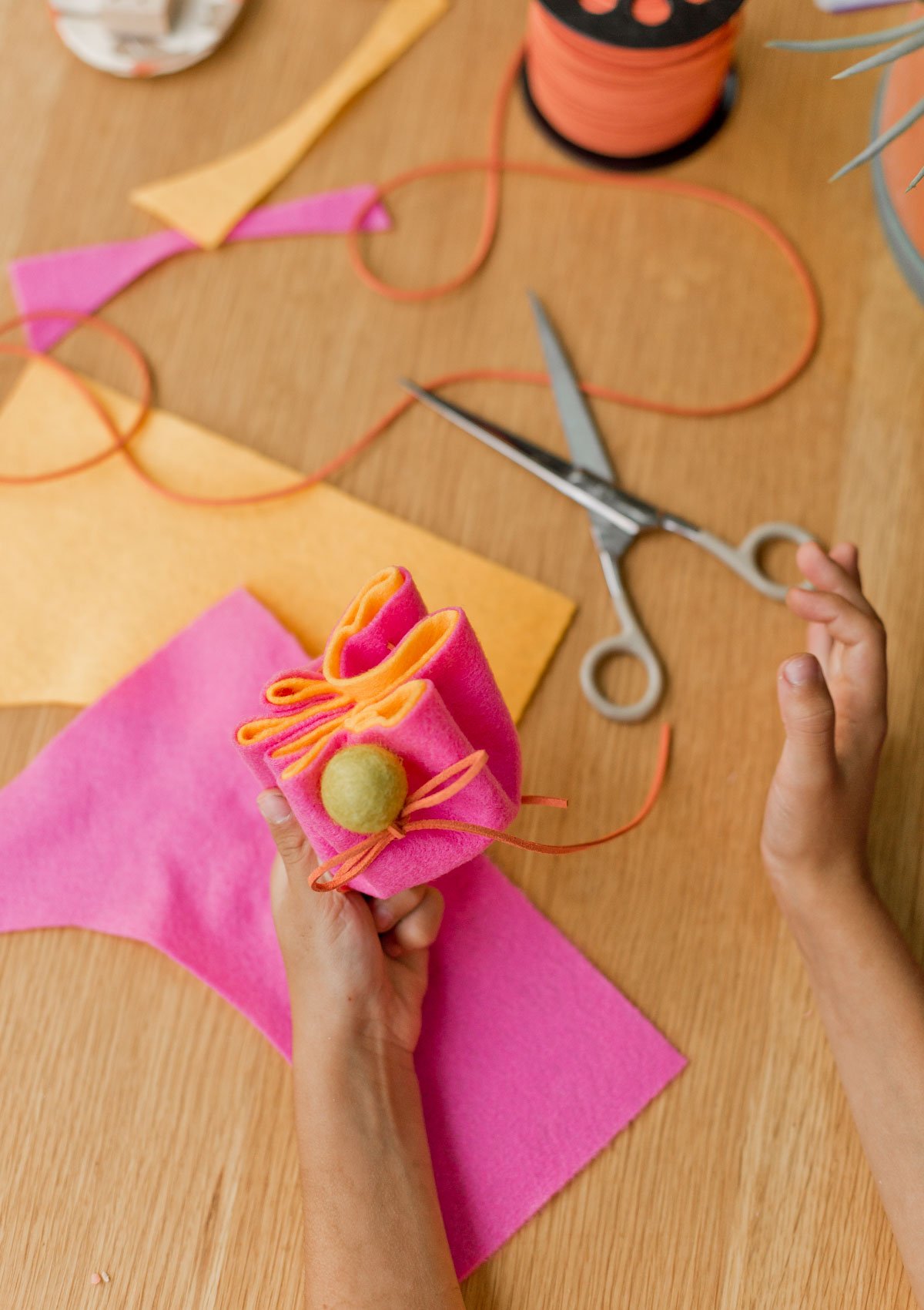 child's hand holding colorful pink and orange felt pouch, with craft supplies all around on the table