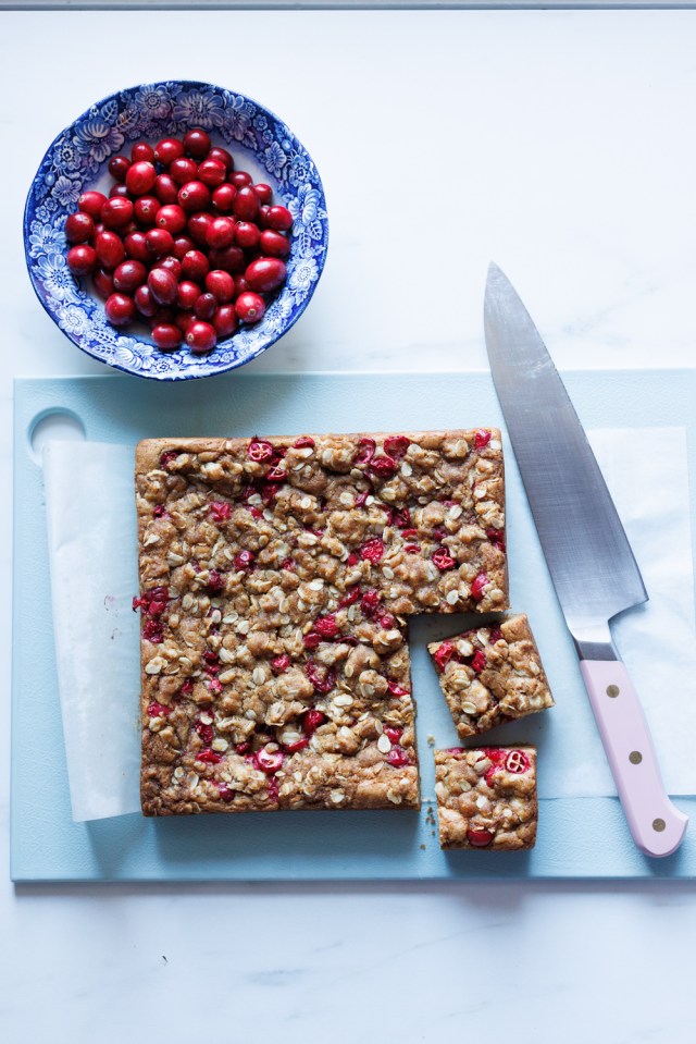 Blondies with cranberries on a blue cutting board next to a chef's knife and a bowl of fresh cranberries.
