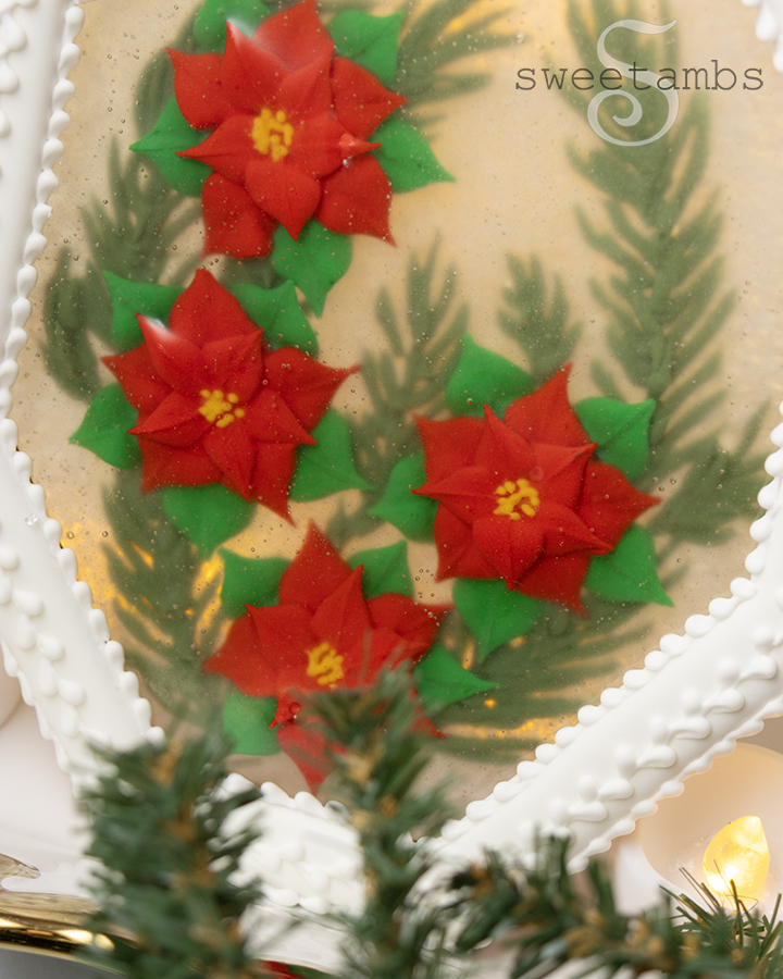 A close up of the edible holiday centerpiece with royal icing poinsettias and royal icing evergreen branches encased in isomalt inside of a white octagon shaped cookie frame covered in royal icing. The centerpiece is in front of a golden bokeh lights background and there are artificial evergreen boughs on the table in front of the centerpiece.