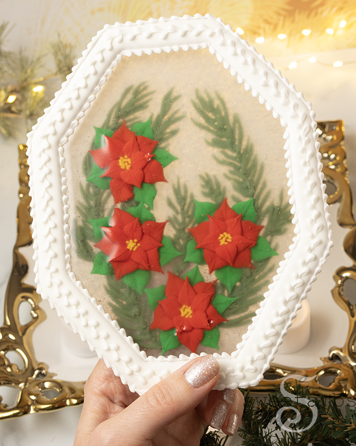 A hand with white shimmery nail polish is holding the edible holiday centerpiece with royal icing poinsettias and royal icing evergreen branches encased in isomalt inside of a white octagon shaped cookie frame. The centerpiece is in front of a gold filigree rimmed platter and fairy lights.