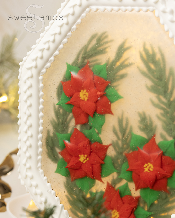 A close up of the edible holiday centerpiece with royal icing poinsettias and royal icing evergreen branches encased in isomalt inside of a white octagon shaped cookie frame covered in royal icing. The centerpiece is in front of a golden bokeh lights background and there are artificial evergreen boughs on the table in front of the centerpiece.